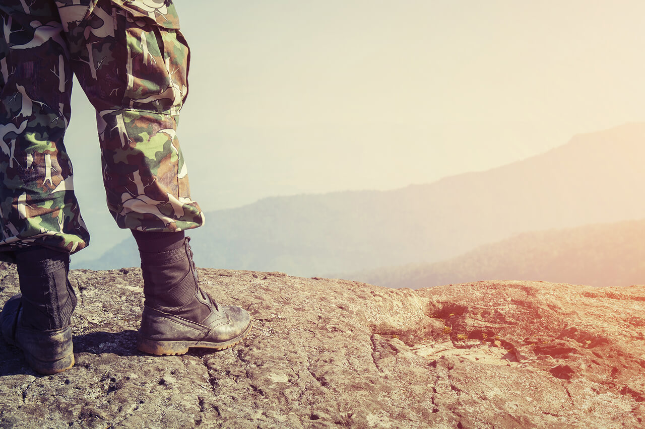 Military soldier standing on a rock outcrop with the sun setting behind, low shot showing feet and legs only